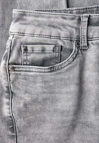 Acercamiento a unos vaqueros de mezclilla gris claro mostrando el bolsillo frontal con un bolsillo pequeño para monedas dentro y con pasadores del cinturón y remaches visibles.