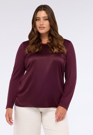 Langärmliges Oberteil in tiefem Burgundy, aus glattem, glänzendem Stoff. Hat einen Rundhalsausschnitt und eine entspannte Passform. Kombiniert mit hellen Hosen.
