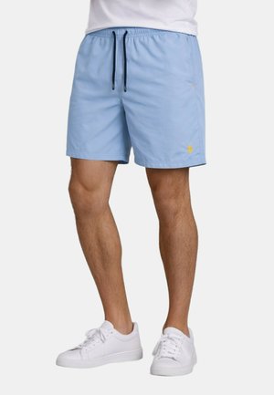 Man wearing light blue drawstring shorts with yellow logo on left leg and white sneakers, standing against plain background.