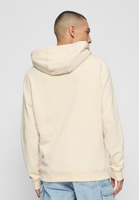 Beige hoodie gemaakt van zachte stof, voorzien van een capuchon met trekkoord, een kangoeroezak en geribbelde manchetten. Achteraanzicht toont een gladde textuur en pasvorm.