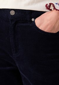 Herren Navy-Corduroy-Hosen aus strukturiertem Stoff mit Fronttasche und Metallknopfverschluss am Bund. Nahaufnahme des Materials und Designs.
