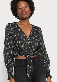 Blusa preta com padrão floral, mangas longas, decote em V, frente com botões, cintura com nó e padrão floral branco sobre um fundo pontilhado.