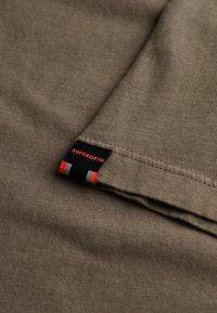 Olijfgroene katoenen stof met een gladde textuur, met een zwart label met rode en witte accenten, gevouwen aan de onderrand.