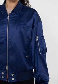 Marineblaue Satin-Bomberjacke mit geripptem Kragen, Reißverschluss vorne, eingesticktem Logo und Reißverschluss-Taschen am Arm. Mit Druckknöpfen am Saum.