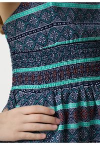 Blauwe jurk met een patroon, voorzien van een geschulpt lijfje en een gerimpelde zoom. De stof bevat geometrische ontwerpen in teal, wit en rode accenten.