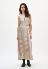 Złota metaliczna sukienka maxi z plisami, dopasowanym gorsetem i wiązaniem w talii, z okrągłym dekoltem i zwiewną spódnicą, zestawiona z czarnymi botkami za kostkę.