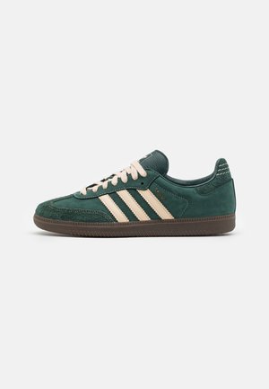 Zielony zamszowy sneaker z beżowymi paskami, beżowymi sznurowadłami i brązową podeszwą, pokazany z boku na białym tle.