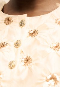 Κοντινή λήψη κρεμ υφάσματος με χρυσή παγιέτα σε λουλουδένια κέντημα και στρογγυλά κουμπιά με στρας στην περιοχή του μπροστινού λαιμού.