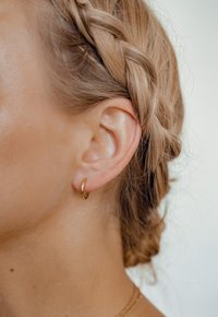 Pendiente de aro dorado exhibido en la oreja de un modelo, acentuado por un peinado trenzado y una textura de piel suave. Diseño elegante y minimalista.