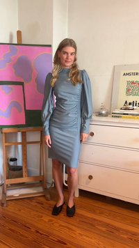 Taivaansininen istuvapuku, jossa on pitkät puff-hihat ja rypytetty sivuyksityiskohta. Mustat neliönpääkengät. Yksinkertainen, teksturoitu kangas.