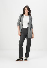 Un long cardigan gris sur un top blanc, associé à un pantalon gris bien taillé et des sneakers noires avec des lacets beige. Design simple, textures variées.
