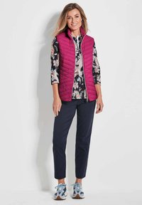 Magenta gesteppte Weste, blumengemustertes Oberteil, marineblaue Hose und lässige blaue Sneaker. Hervorzuhebende Merkmale sind der Reißverschluss und die verstellbare Passform.