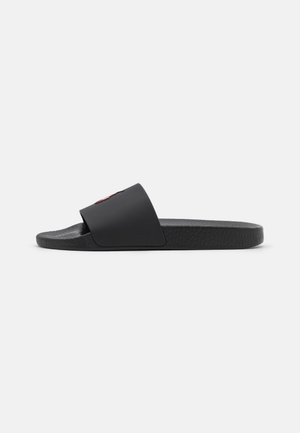 Sandale slide noire avec semelle texturée et large bride sur le dessus affichant un petit logo rouge, vue de côté sur fond blanc.
