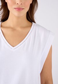 T-shirt bianco in cotone con scollatura a V e leggero dettaglio sulle spalle. Presenta una texture liscia e dettagli in pizzo lungo il colletto.