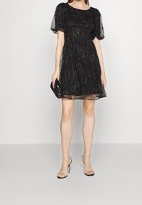 Robe noire avec des manches courtes et transparentes en forme de volants, ornée de motifs floraux en fil métallique. Assortie d'une pochette noire et de sandales à talons.