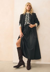 Blazer nero sopra un maglione a motivo, abbinato a una gonna nera fluente e stivali marroni in suede fino al ginocchio. Texture lisce e design a strati.