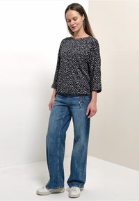 Femme portant un haut à manches longues à motifs noir et blanc, un jean bleu à jambes larges et des baskets blanches, debout devant un fond uni.