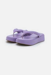 Violetti alusta varvasvarvashapsut pehmustetuilla remmeillä ja ruskeilla tasapohjilla valkoista taustaa vasten, kulmassa esitettynä etu- ja sivunäkymissä.