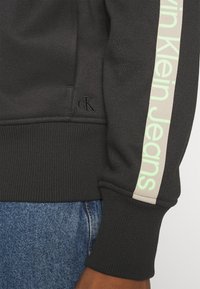 Felpa nera con polsini a costine, caratterizzata da un logo "Calvin Klein Jeans" verde chiaro su una striscia crema. Include un piccolo logo in rilievo.