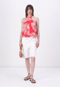 Red halter top with white pattern, white shorts, brown sandals, and a small brown handbag. Model stands against a plain background.