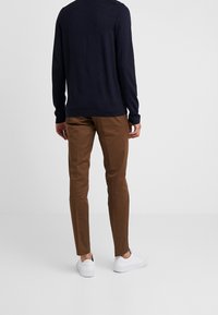 Marinblå långärmad skjorta, bruna slim-fit byxor och vita sneakers ses bakifrån. Tyget verkar slätt med standardstickning.