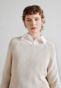 Lys beige ribbet genser med vid halsringning og lange ermer, lagd over en klar hvit skjorte med krage. Subtil tekstur og avslappet passform.