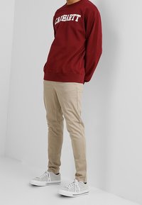 Bordeaux crewneck sweatshirt met witte "CARHARTT" logo, gecombineerd met beige slim-fit broek en grijze sneakers met witte veters.