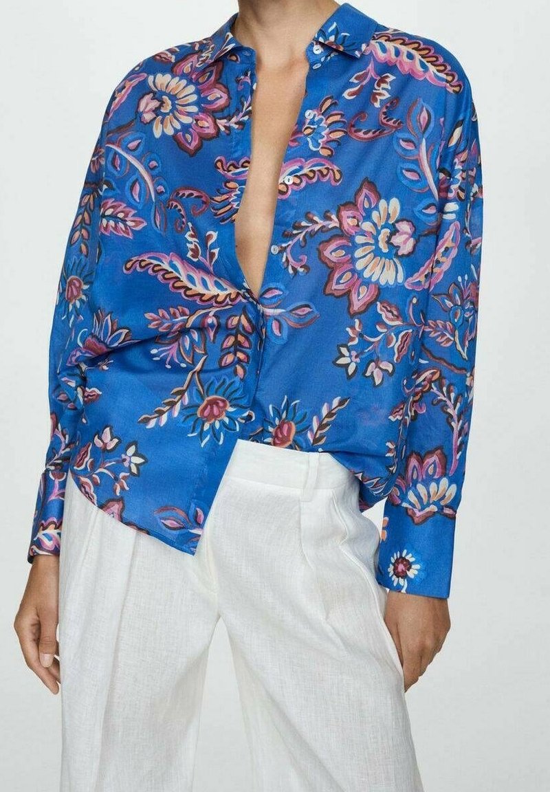 Personne portant une chemise à fleurs bleu vif à boutons, partiellement déboutonnée, rentrée dans un pantalon blanc ample, debout devant un fond uni.