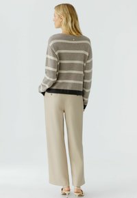 Vrouw die van zich af kijkt, gekleed in beige wijde broek, een taupe en grijs gestreepte trui, en witte hakken tegen een effen achtergrond.