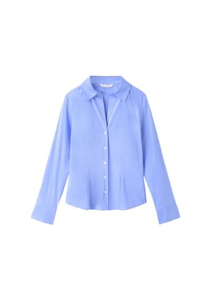 Jeune femme portant une chemise bleue déboutonnée, un jean à jambes larges avec des revers, des chaussures pointues noires et un vêtement noir noué à la taille.