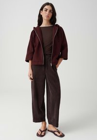 Woman standing wearing a maroon zip-up jacket, dark wide-leg pants, and maroon sandals against a plain light background.