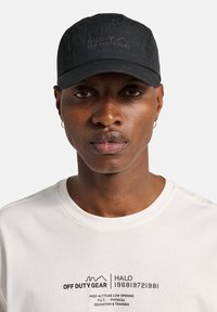 Czarna czapka z fakturowanej tkaniny z wypukłymi literami; noszona z białym t-shirtem z czarnym tekstem i liniami graficznymi.