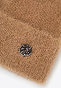 Hnědá pletená čepice s měkkou texturou. Má kulaté kovové logo s hvězdovým motivem. Materiál má teplý, chlupatý vzhled.