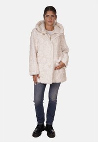 Giacca in pelliccia finta color crema, con cappuccio, chiusura con zip e tasche frontali; abbinata a jeans blu e stivali neri. Tessuto morbido e vestibilità ampia.
