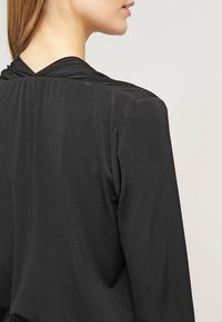 Vêtement noir à manches longues avec un design de dos drapé et une texture lisse. Il présente un décolleté en V profond et une coupe décontractée.
