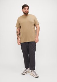 Tan t-skjorte med kort erm og hvit logo, kombinert med mørke cargo-bukser og grå joggesko, med en avslappet passform og glatt tekstur.