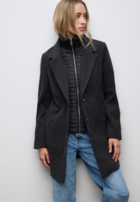 Sort uldblandet frakke med en lang silhuet, brede klapder og to knapper, lagdelt over en sort quiltet jakke, parret med blå jeans.