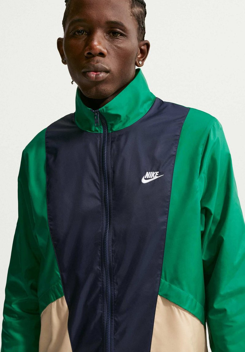 Junger Mann trägt eine grüne, marineblaue und beige Nike Jacke mit Reißverschluss, hohem Kragen und sichtbarem Logo vorne.