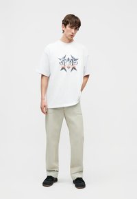 Noor mees kannab üle suurte valgete graafiliste motiividega t-särki, beežikaid laiade säärtega pükse ja musti kingi, seisab ühe käega taskus.