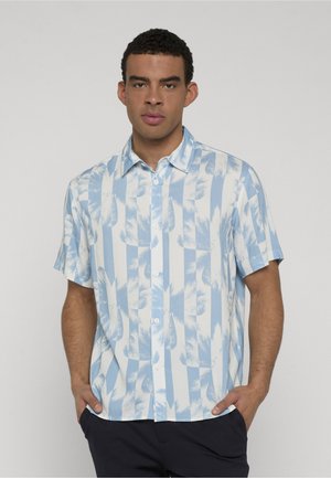 Ung mand iført en blå og hvid kortærmet skjorte med knapper og et palmebladsmønster samt mørke bukser, hænderne i lommerne, foran en ensfarvet baggrund.
