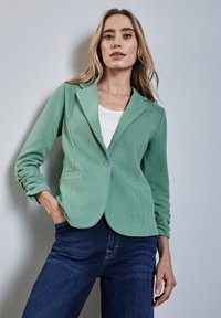 Muntgroene blazer met een getailleerde pasvorm, voorzien van een enkele knoopsluiting, twee voorzakken en gerimpelde mouwen. Draag het over een witte top.