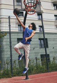 Athlete in a blue sleeveless jersey and white pants jumps to dunk a basketball into an orange hoop. The court is red, with a wire fence background.