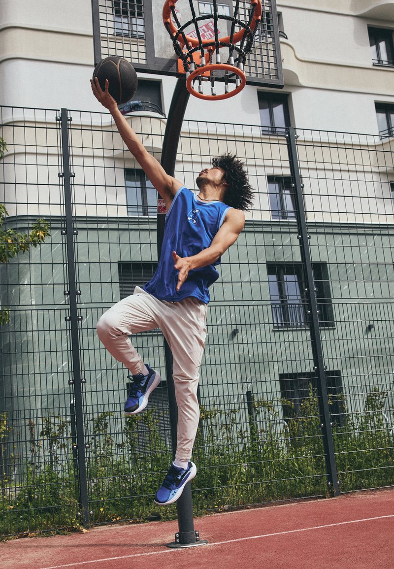 Un athlète en maillot sans manches bleu et pantalon blanc saute pour dunker un ballon de basket dans un panier orange. Le terrain est rouge, avec une clôture en fil métallique en arrière-plan.