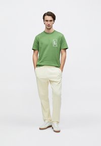 Πράσινο βαμβακερό μπλουζάκι με λευκό γραφικό σχέδιο στο στήθος, συνδυασμένο με μπεζ παντελόνι και λευκά αθλητικά παπούτσια με καφέ σόλες.