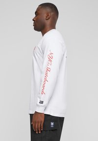 Long-sleeve white cotton shirt featuring red text on the left sleeve and a logo patch at the hem. The design is simple and straight-cut.