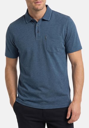 Homme portant un polo bleu à manches courtes avec une poche avant et un pantalon foncé, les mains détendues le long du corps.