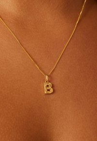 Collier en or avec un pendentif texturé en forme de grande lettre "B". La chaîne est fine et élégante, reposant sur une peau lisse.