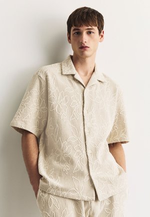 Noor mees kannab kreemjat tekstuurset lühikese varrukaga särk ja sobivaid pükse õrnade lillemustritega, seisab ühevärvilise heleda seina ees.