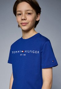 Modrá bavlněná tričko s bílým logem "TOMMY HILFIGER EST. 1985" a malou vlajkovou akcentací na rukávu. Krátké rukávy, kulatý výstřih.
