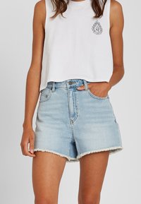 Top corto blanco con un pequeño diseño gráfico negro en el lado izquierdo del pecho, combinado con pantalones cortos de mezclilla de talle alto en azul claro con dobladillos desgastados.